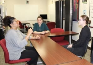 Three students sit around a table in a classroom practicing signing. One student is actively signing and the other two are watching.