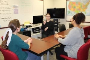 Three students sit around a table in a classroom learning signs. One student sits in the center of the frame facing the camera and the other two sit on either side facing away. The center student is showing a sign and the other two are watching.
