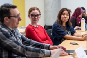 An image of a classroom. A man in a plaid shirt and glasses is looking at an Ipad, and two female classmates, one with glasses and a red shirt and one with long dark hair and a blue shirt are looking and appear to be listening.